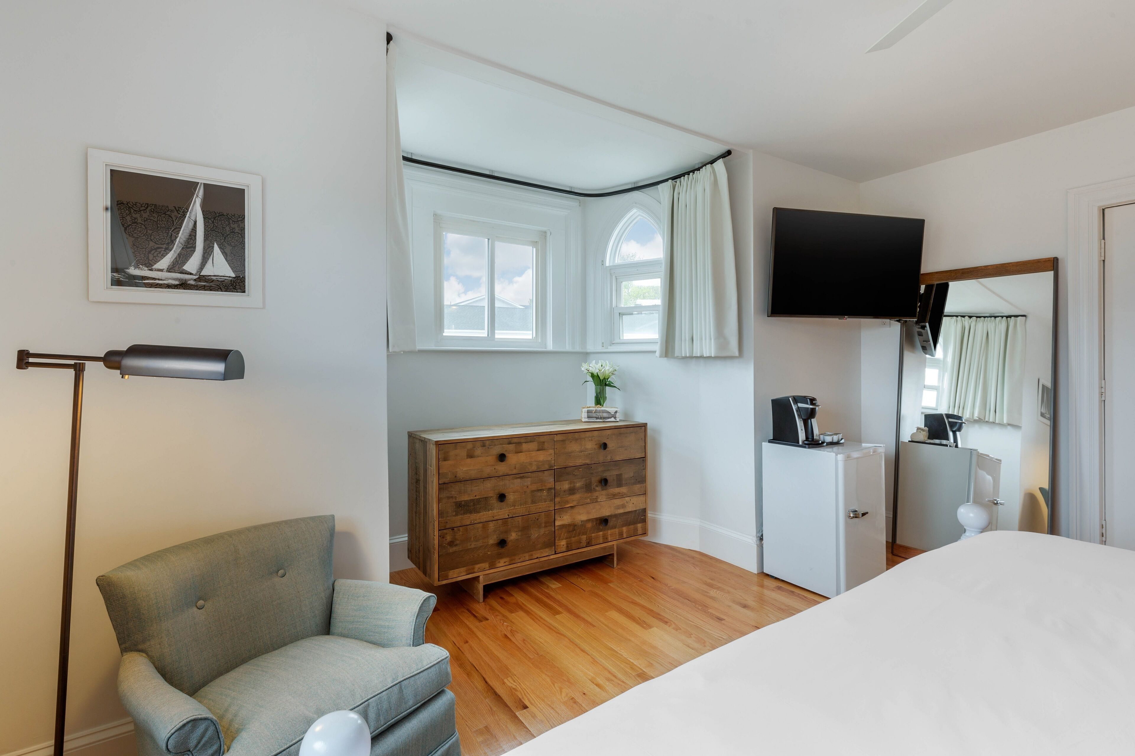 Un dormitorio con cama, cómoda de madera, televisor, silla y ventana con cortinas.