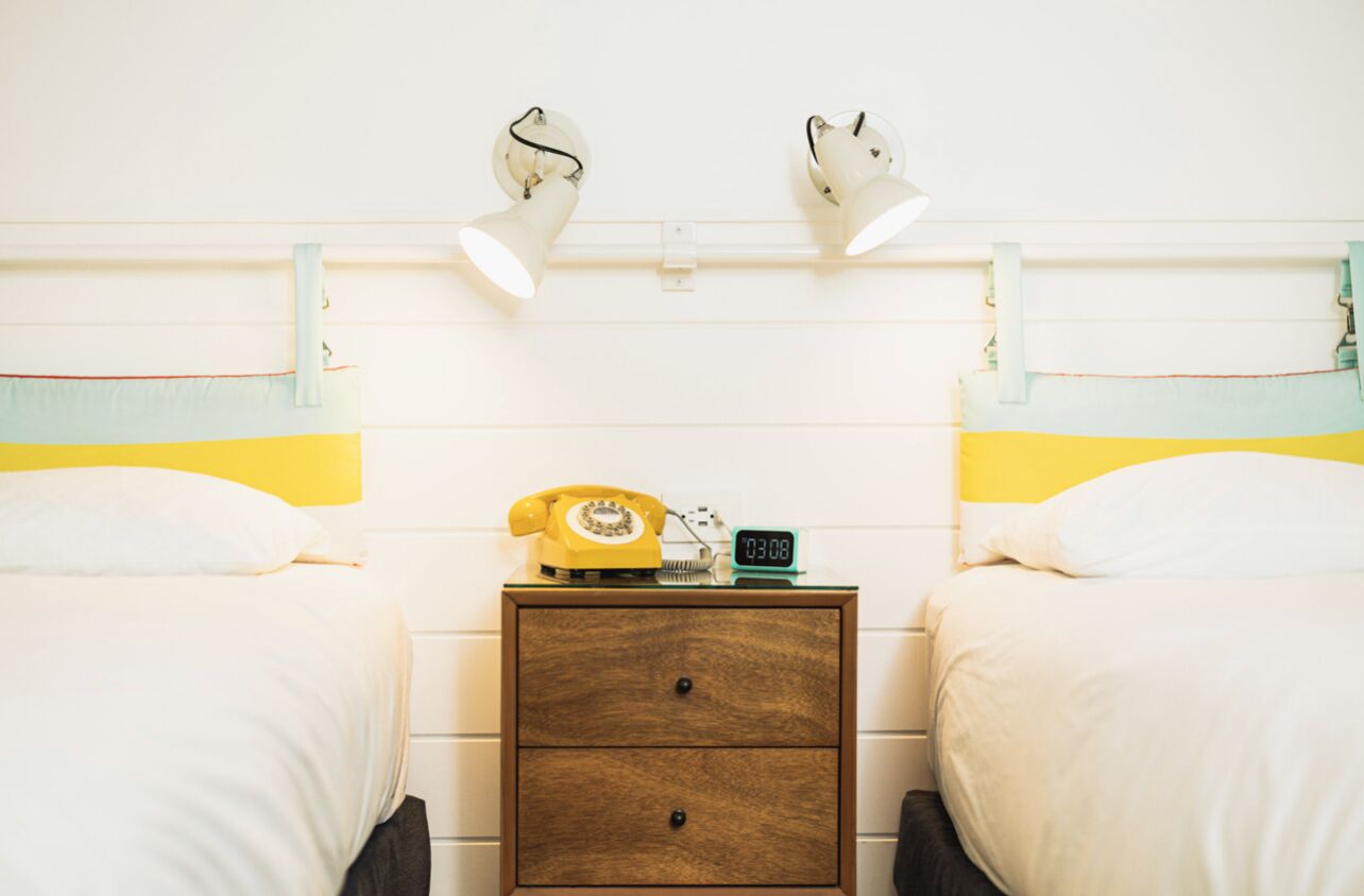 Een tweepersoonskamer met twee bedden, een houten nachtkastje, een gele draaischijf telefoon en een digitale klok.