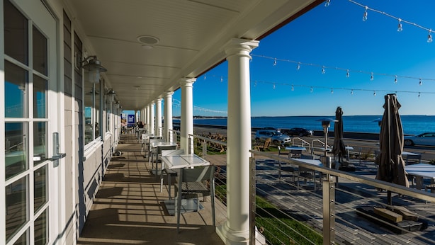 A covered outdoor area with tables and chairs, a view of the sea, and a building with large windows. - The Shore House (Narragansett)