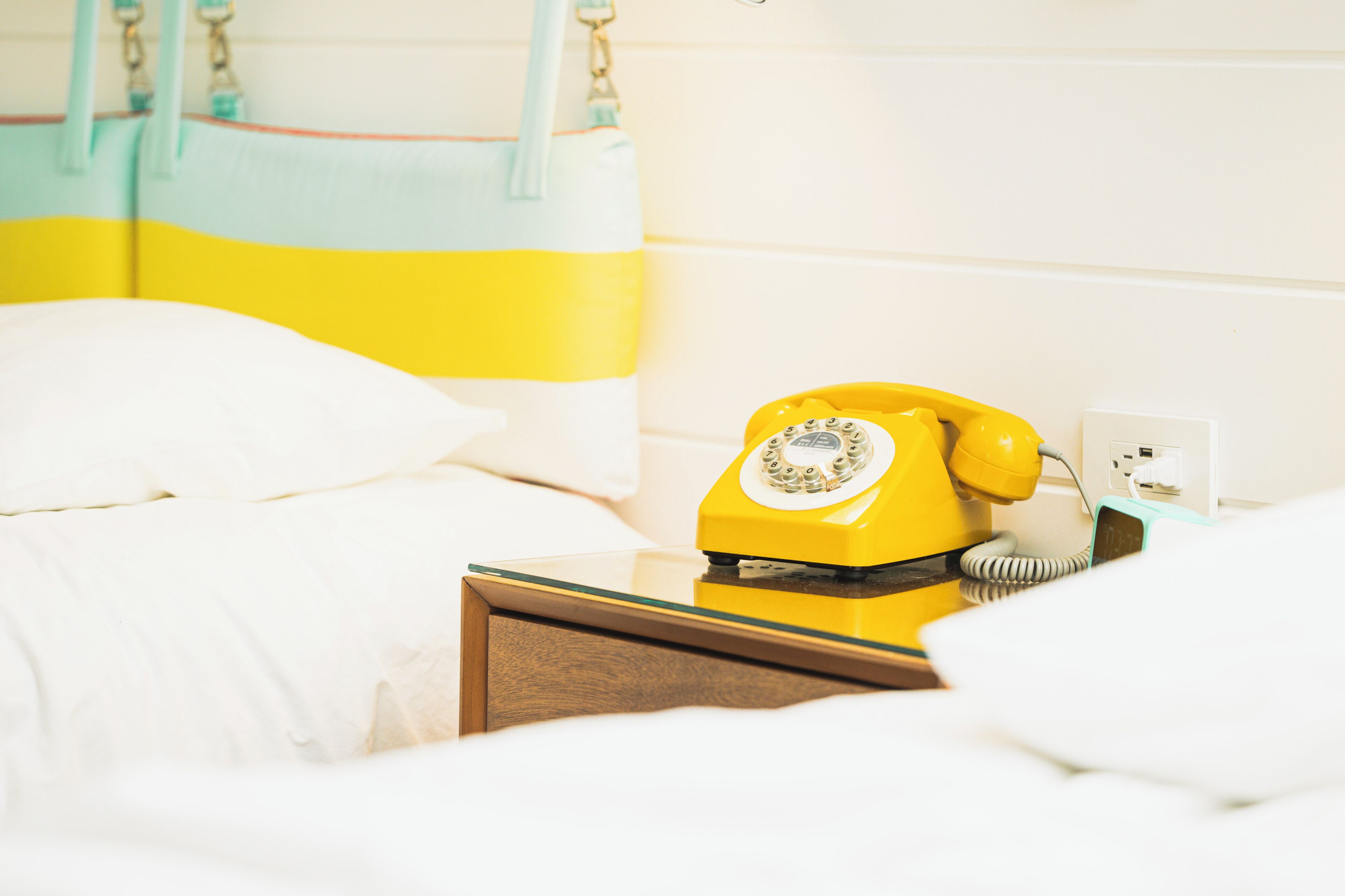 A bed with white bedding and a yellow rotary phone on a bedside table.