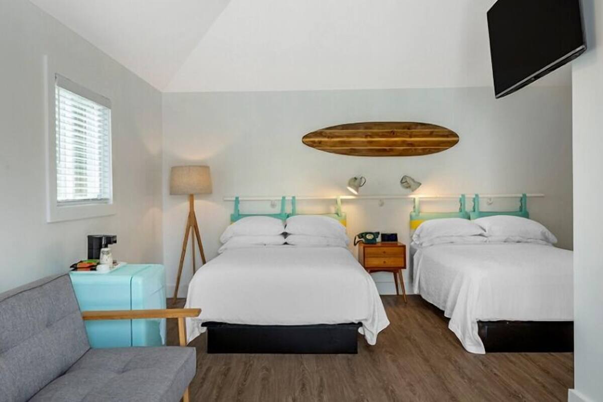 A hotel room with two beds, a sofa, a lamp, a TV, and a window with blinds.