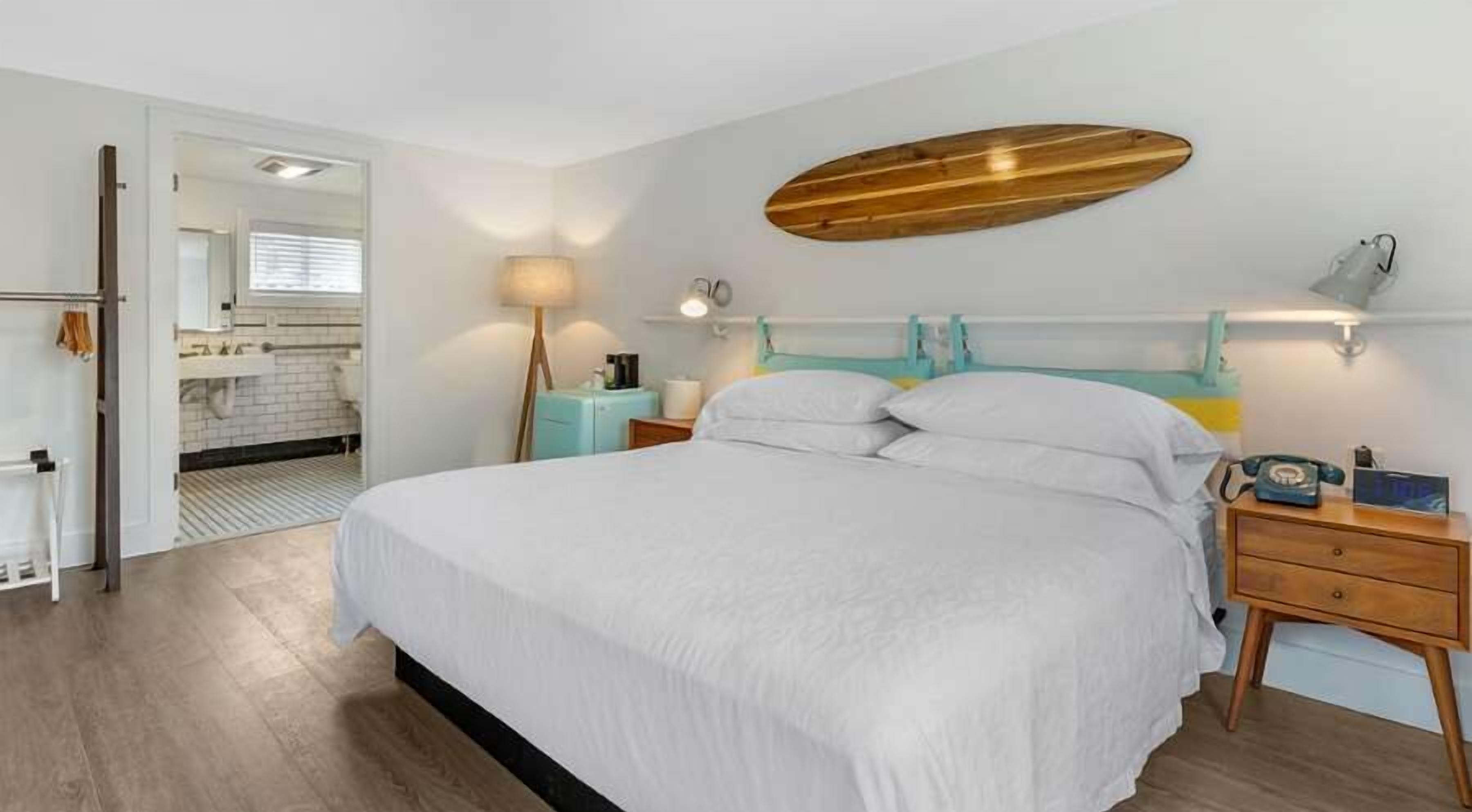 Une chambre à coucher avec un grand lit, des tables de chevet, une étagère fixée au mur et une salle de bain visible à travers une porte ouverte.