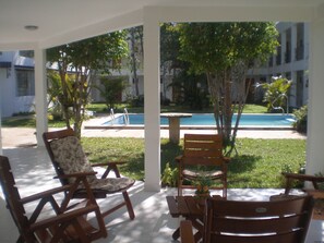 A covered outdoor seating area with wooden chairs and a table, overlooking a swimming pool and greenery.