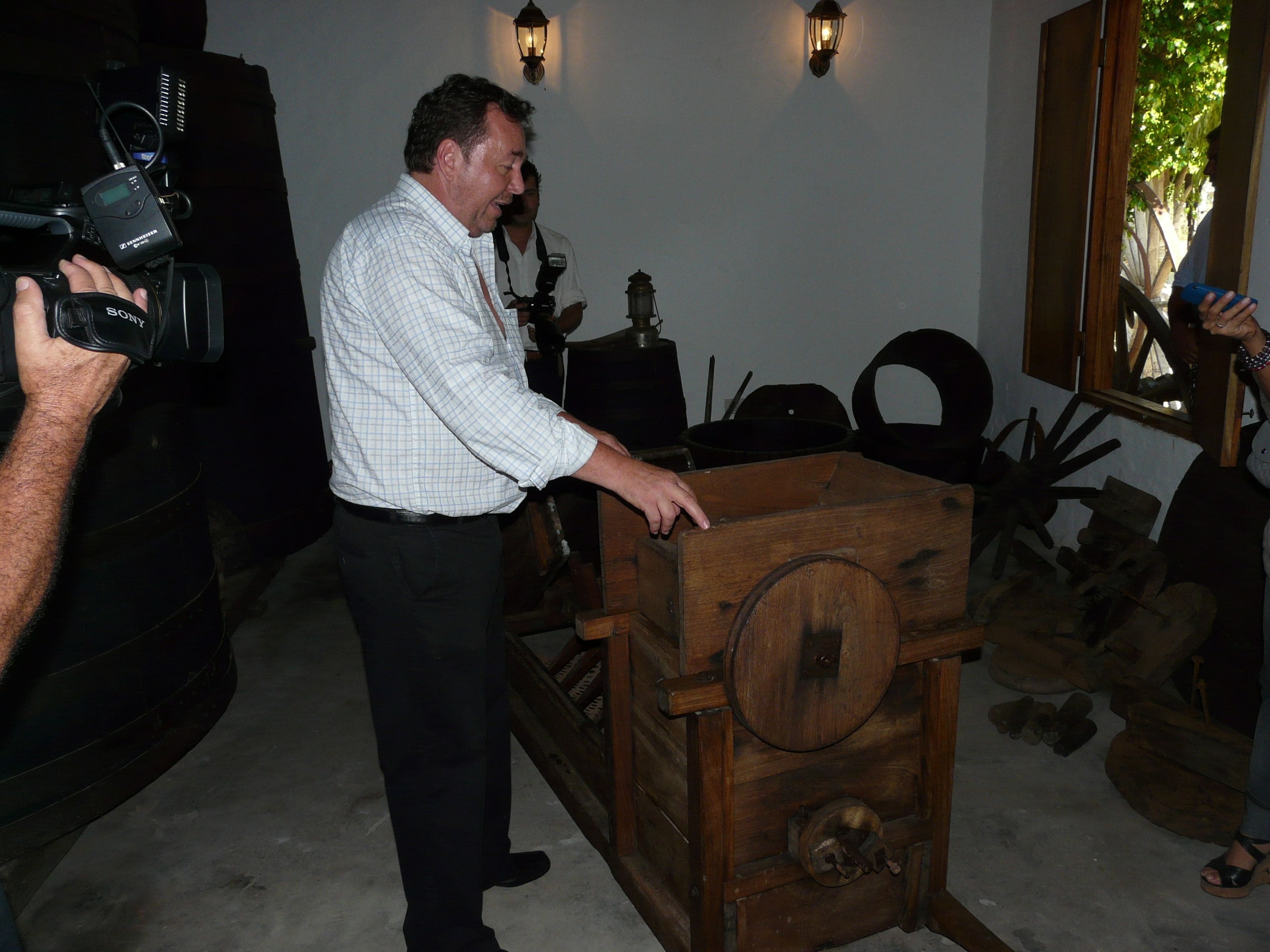 Un hombre de pie junto a un cofre de madera en una habitación con piso de concreto, una mesa de madera y una silla.