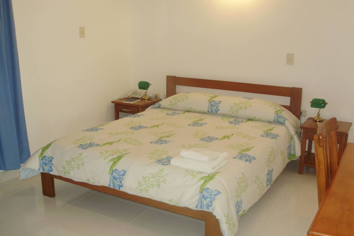 A neatly made bed with floral bedding, a bedside table with a lamp, and a wooden chair.