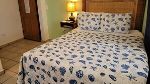 A hotel room with a bed featuring a sea-themed bedspread, a wooden door, a nightstand with a lamp, and a framed artwork on the wall.