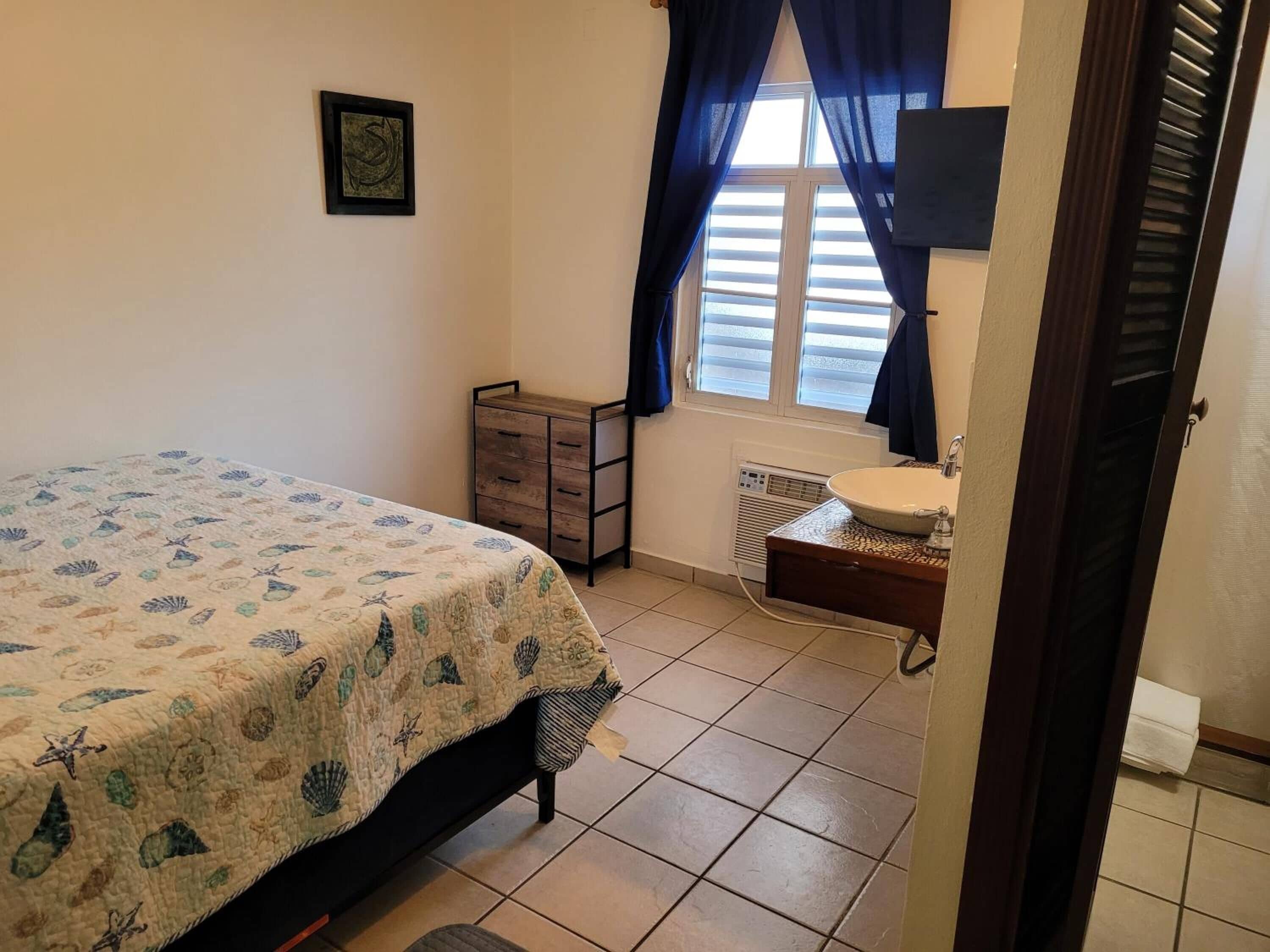 Un dormitorio con cama, cómoda, ventana con cortinas, televisor montado en la pared y radiador.