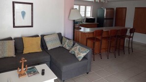 A compact living space with a grey sofa, a small coffee table, and a kitchen area with a fridge, microwave, and bar stools.