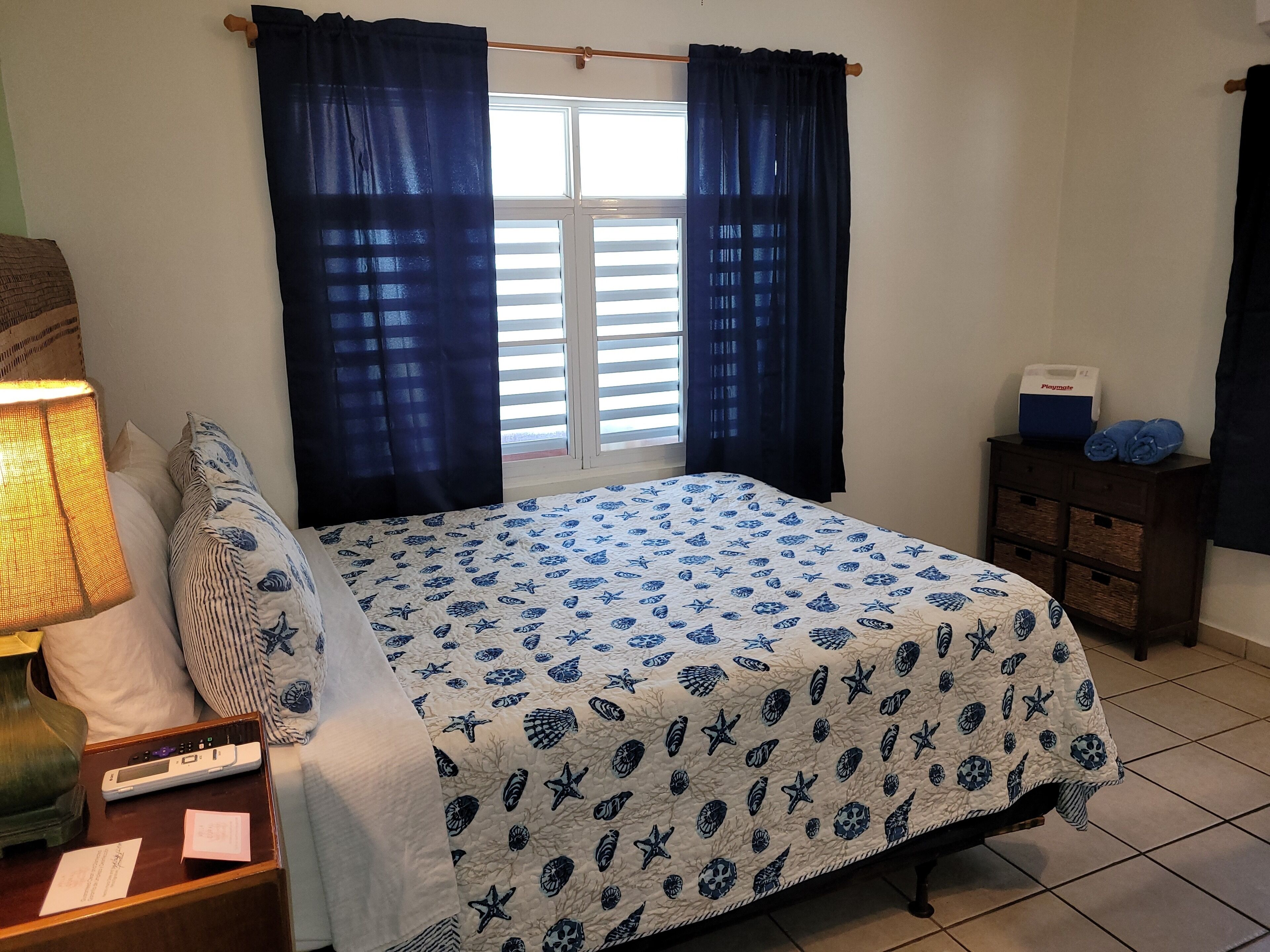 Un dormitorio con cama, cortinas, mesita de noche con lámpara, cómoda y ventana con persianas.