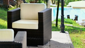 Una silla de mimbre con cojín bajo una pérgola, rodeada de follaje y una piscina al fondo.