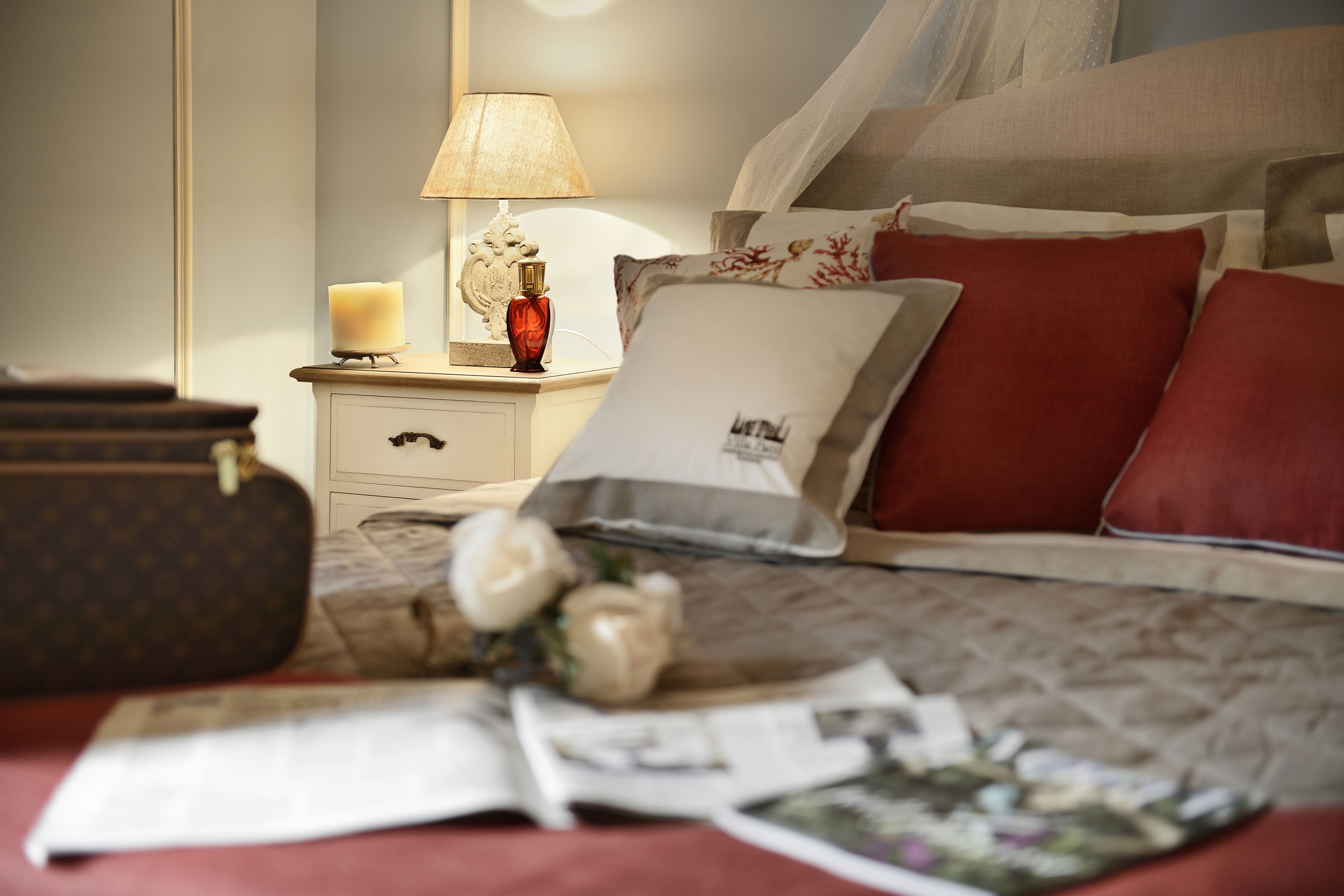 Una cama bien hecha con almohadas rojas y blancas, una lámpara de noche y un jarrón decorativo con flores.