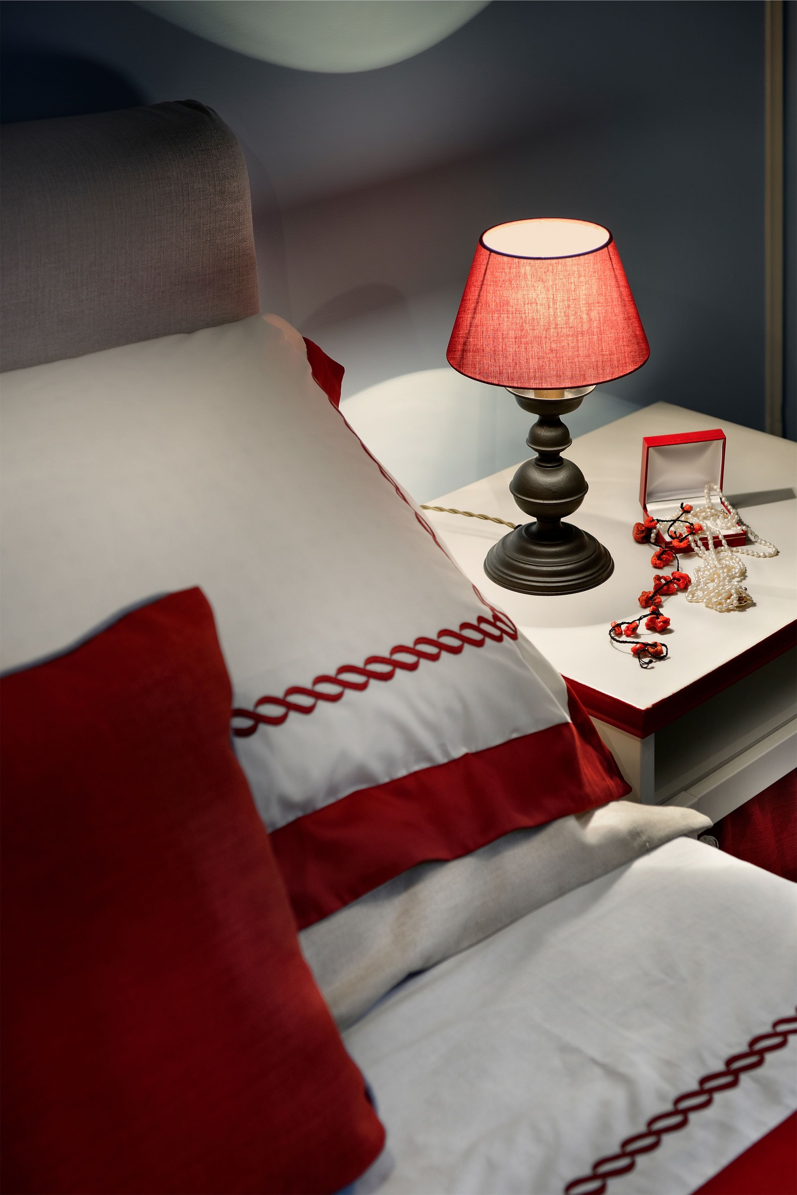 Ein ordentlich bezogenes Bett mit roter und weißer Bettwäsche, ein Nachttisch mit Lampe und eine kleine rote Schachtel.