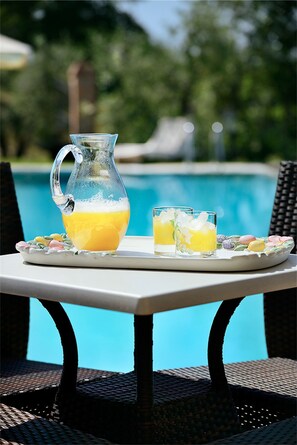 A table with a pitcher of orange juice and two glasses, a plate with colorful macarons, and a view of a swimming pool. - Villa Parri (Pistoia)