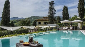 Zona de piscina con sillones de playa y sombrillas, rodeada de vegetación y un edificio con techo de tejas.