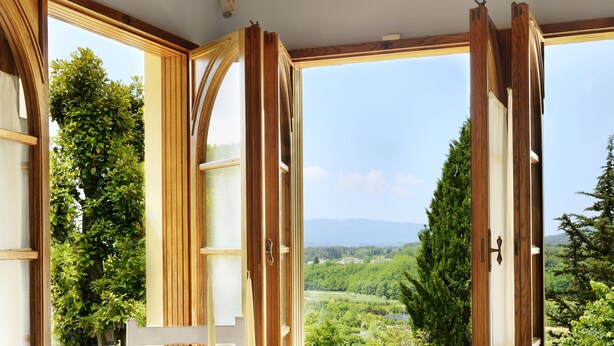 Un comedor con una mesa dispuesta para dos, sillas de madera y vistas a un paisaje verde a través de ventanas abiertas.