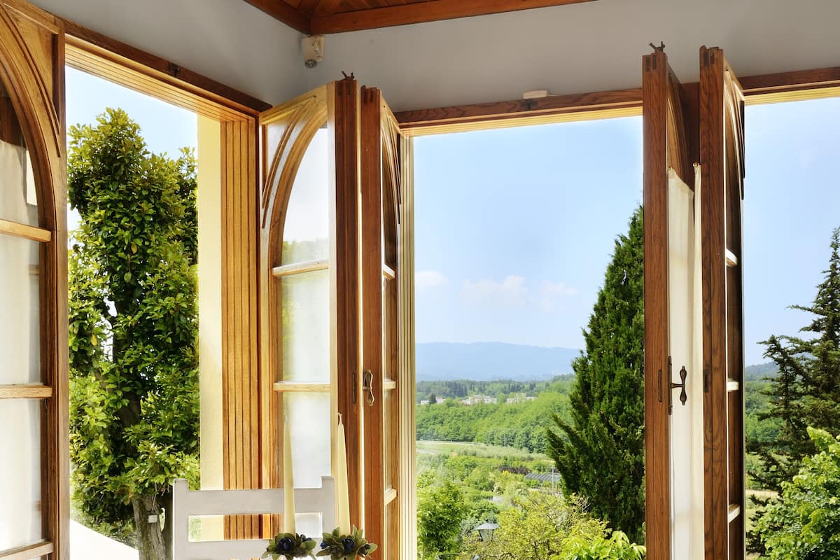 A dining area with a table set for two, wooden chairs, and a view of a green landscape through open windows.