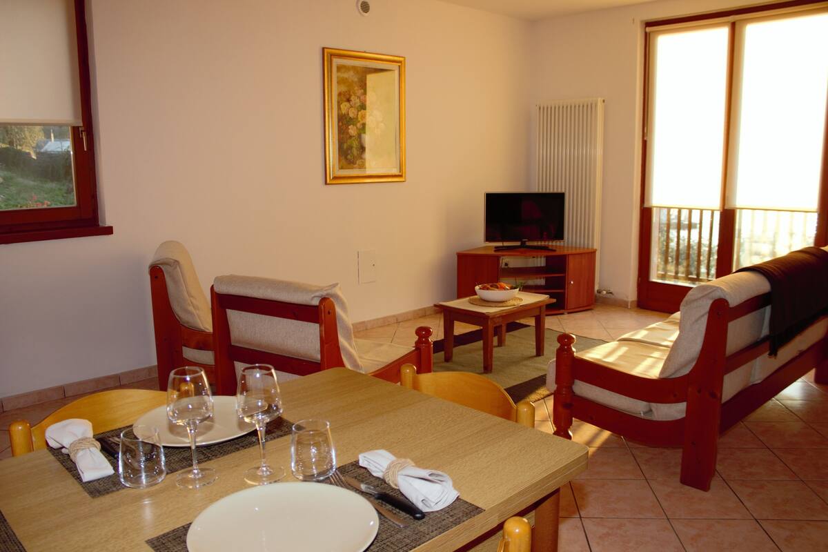 Un comedor con una mesa lista para dos personas, un sofá, un televisor y una ventana con vistas.