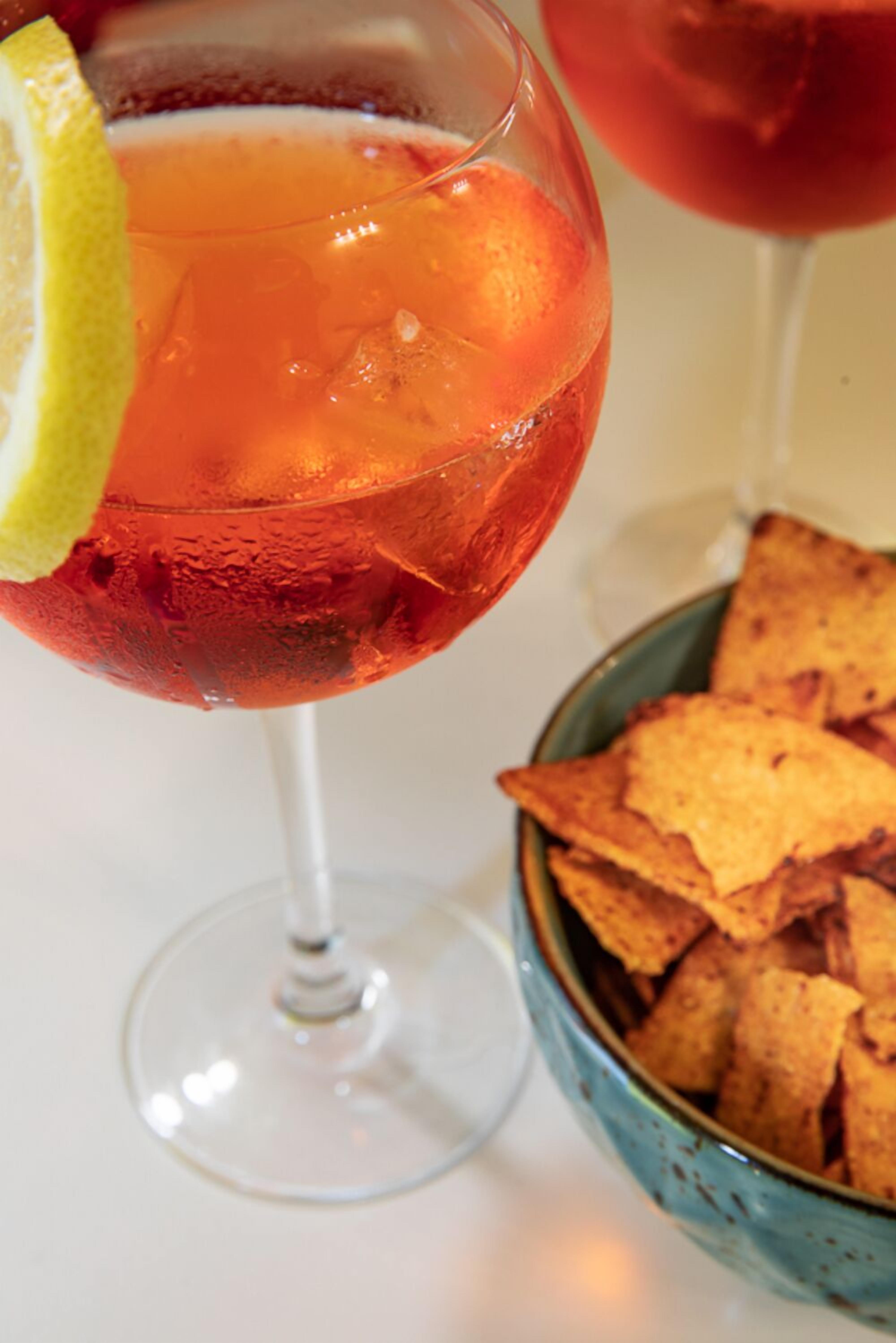 Ein Cocktailglas mit Zitronenscheibe und Eiswürfeln, sowie eine Schale Tortilla-Chips im Hintergrund.