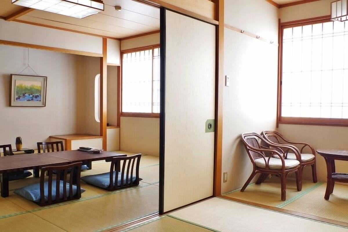 Una habitación japonesa tradicional con suelo de tatami, una mesa baja con sillas, una puerta corrediza y un ventanal con paneles shoji.