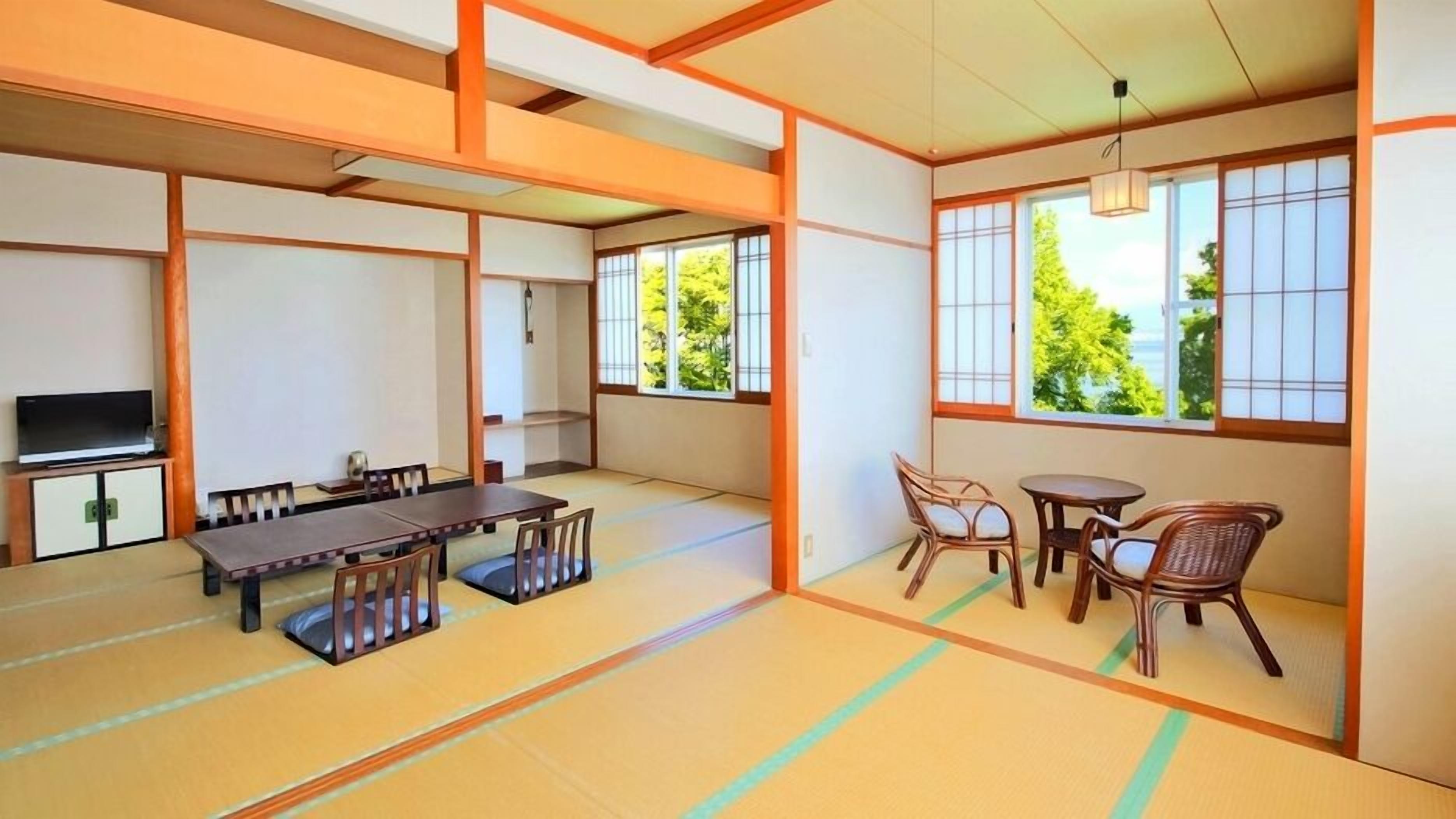 A traditional Japanese room with tatami flooring, a low table with chairs, a television, and large windows with a view of greenery.