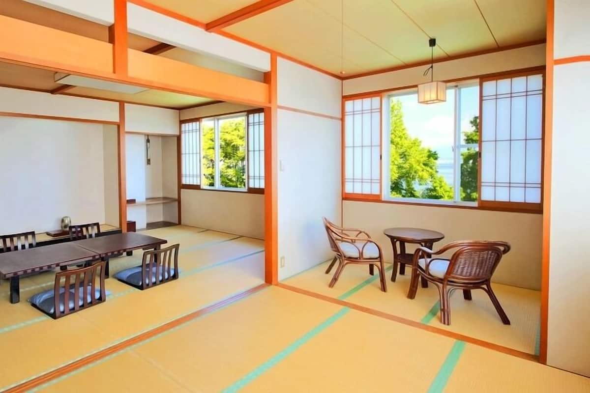 Una habitación tradicional japonesa con suelo de tatami, una mesa baja con sillas, un televisor y ventanas grandes con vistas a la vegetación.