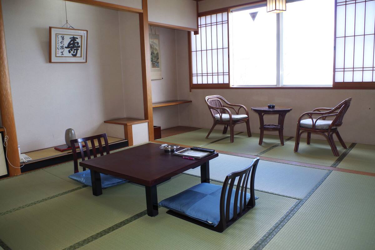 Una habitación japonesa tradicional con suelo de tatami, una mesa baja con cojines y sillas de madera.