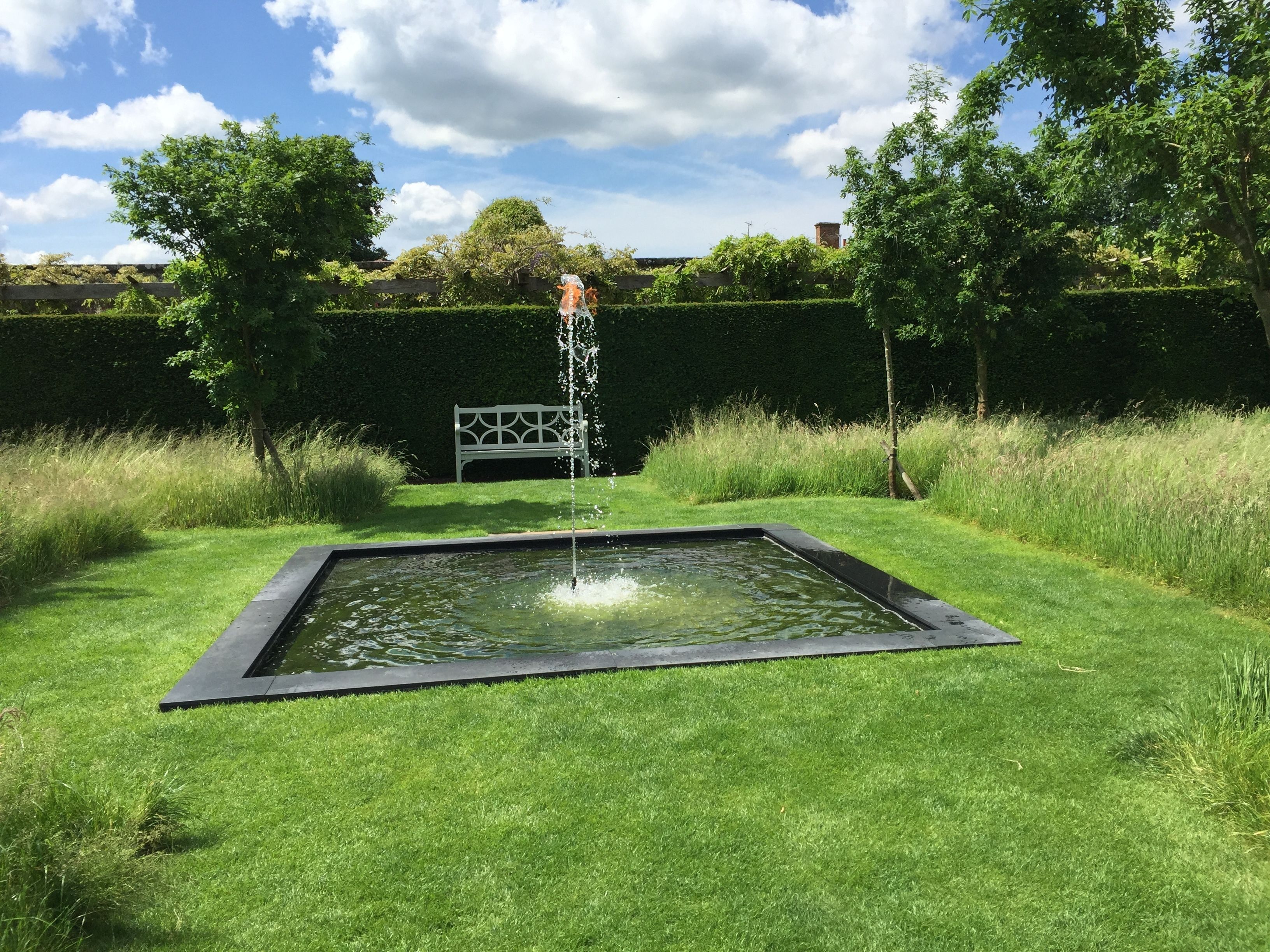 A rectangular pond with a fountain in the center, surrounded by well-maintained grass and a hedge.