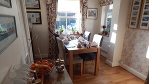 A dining area with a wooden table, chairs, and a fruit bowl. There are framed pictures on the walls and a window with curtains. - Fairlight Lodge (King's Lynn)