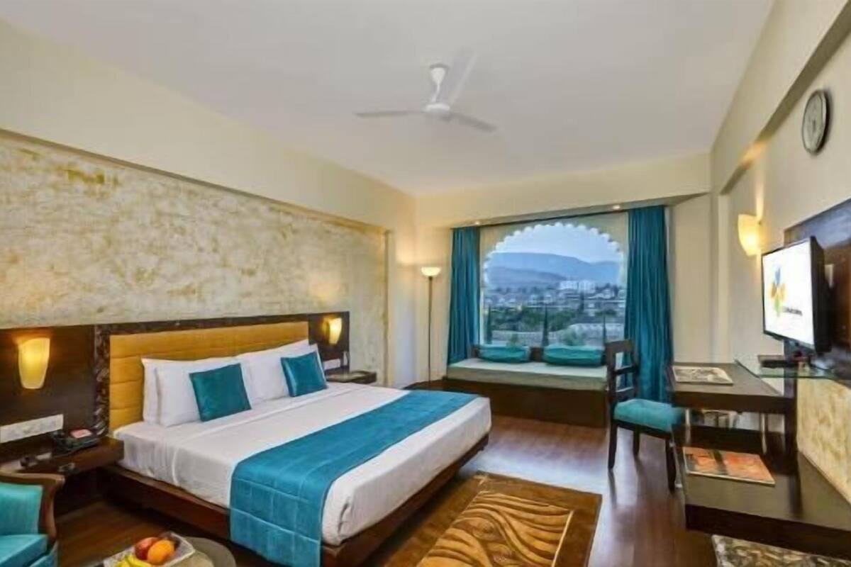A hotel room with a bed, a desk, a chair, a television, and a view of a cityscape.