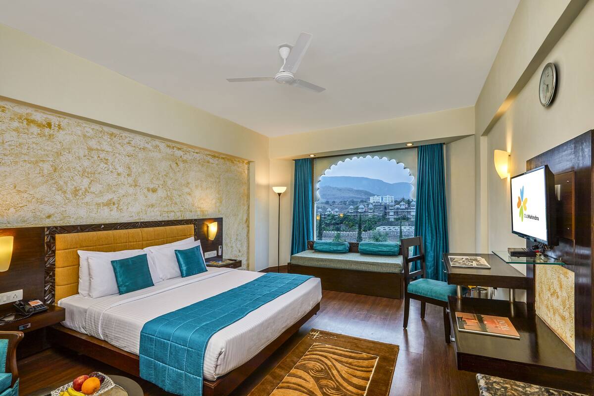A hotel room with a large bed, a desk, a television, and a window with a city view.