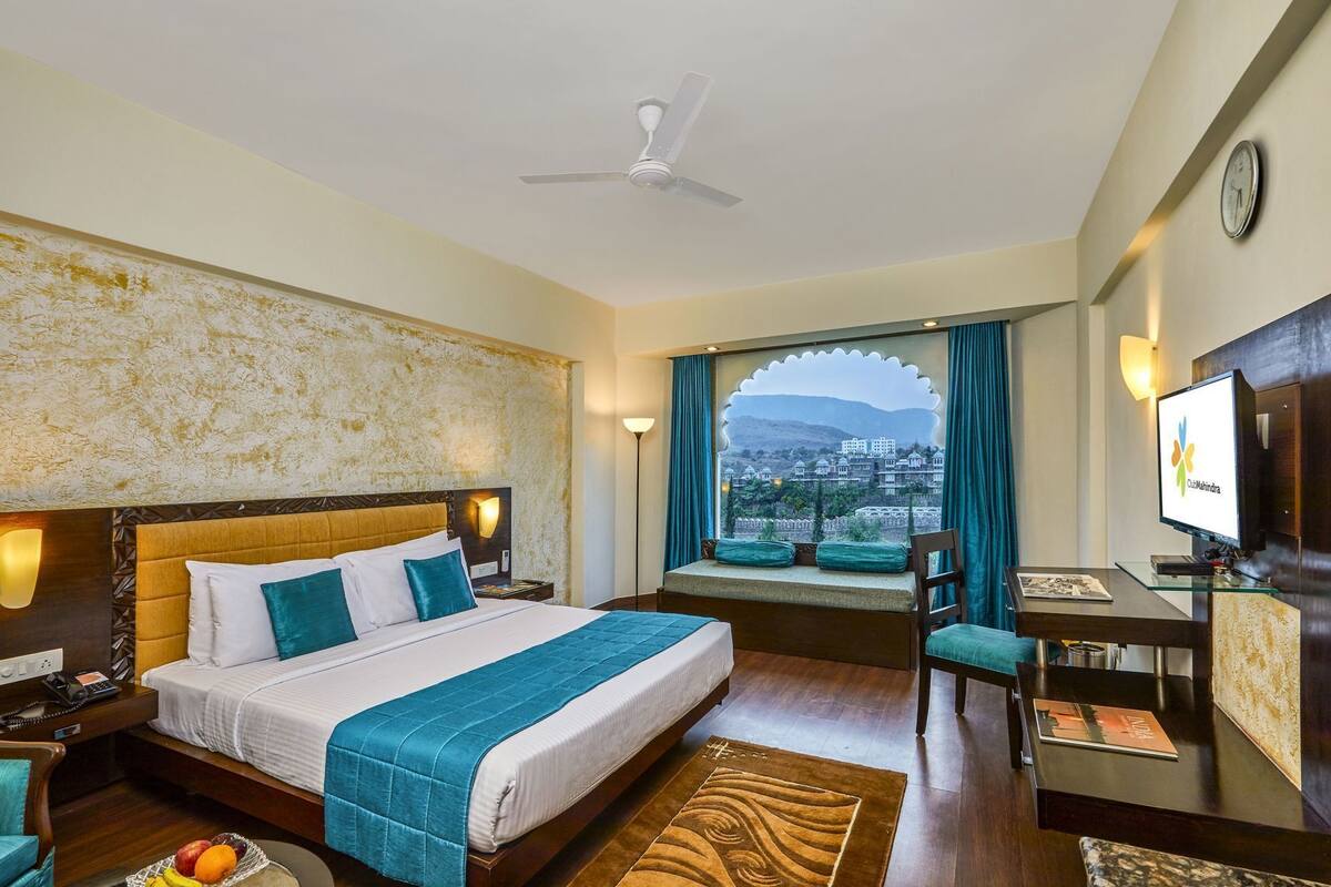 A hotel room with a bed, a desk, a chair, a TV, a clock, and a view of a cityscape.