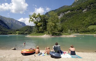 Escena junto al lago con gente relajándose en la orilla, un bote y una vista despejada de las montañas circundantes.