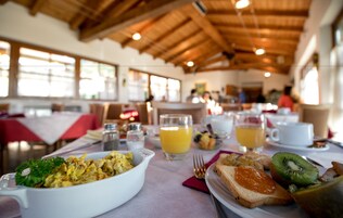 Un desayuno con un plato ovalado blanco de huevos revueltos, un vaso de jugo de naranja, un plato con tostadas y kiwi, y una taza blanca sobre una mesa con una servilleta roja.