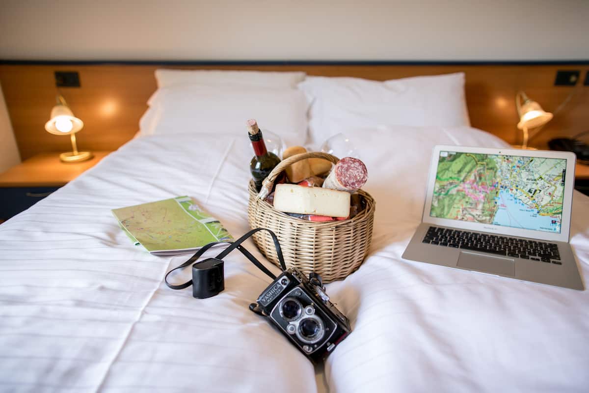 A neatly made bed with a laptop displaying a map, a camera, and a basket of snacks.