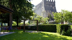 A castle with a stone wall and towers, surrounded by a well-maintained garden with a wooden pergola and swings. - Hotel Antica Croce (Tenno)