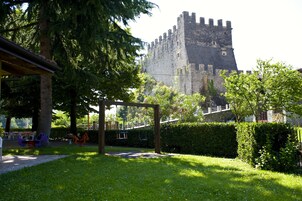 Un castillo con muro de piedra y torres, rodeado de un jardín bien cuidado con una pérgola de madera y columpios.