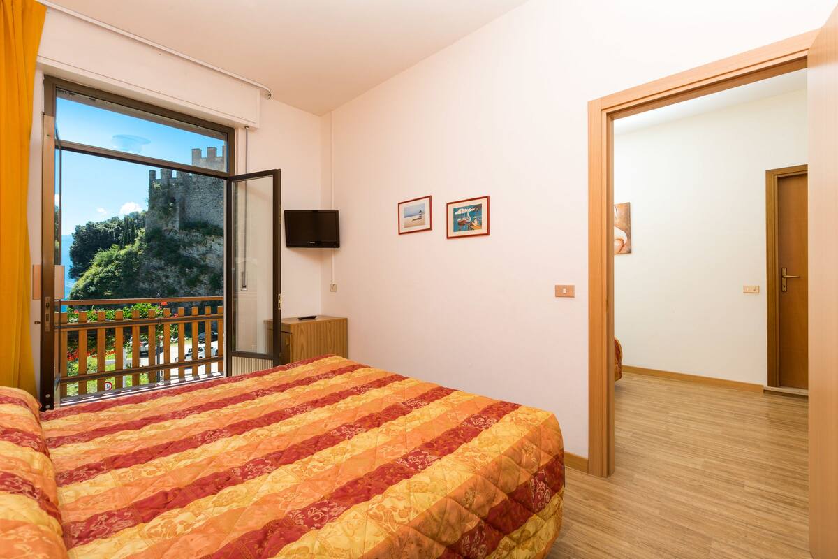 A hotel room with a large bed, a TV, and a balcony with a view of a castle and greenery.