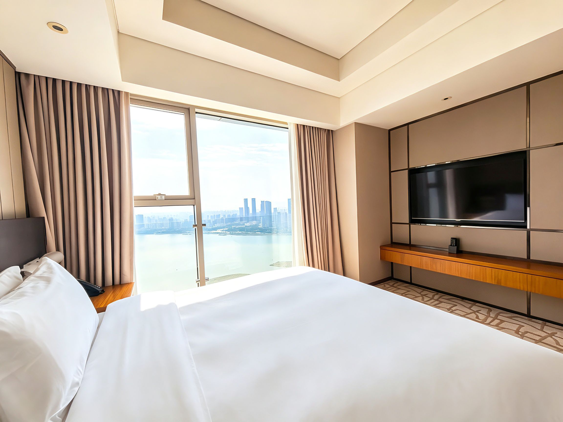 A hotel room with a large bed, a TV on a wooden console, and a view of a cityscape through a large window.