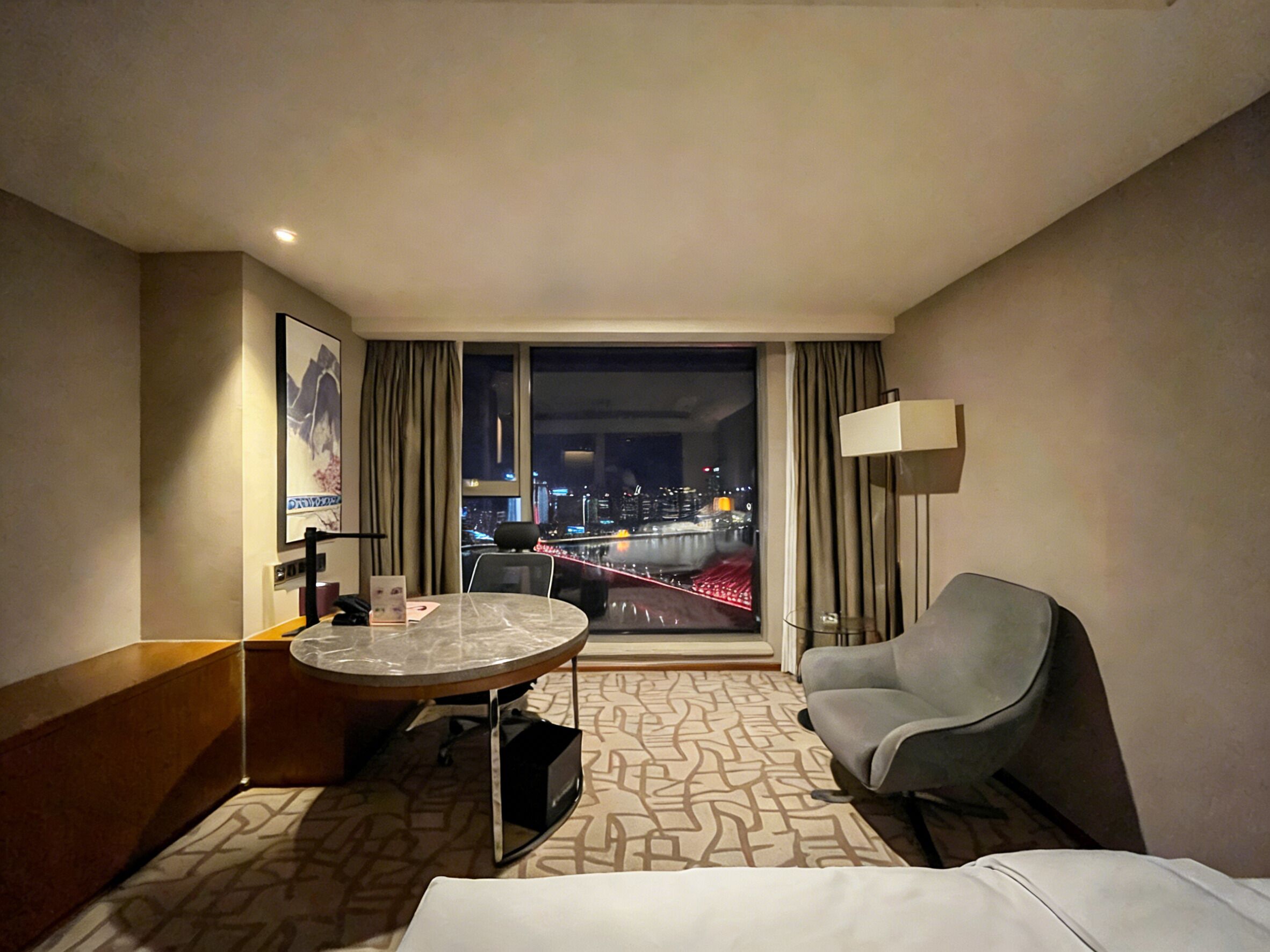 A hotel room with a bed, a desk, a chair, a window with a city view, and a patterned carpet.