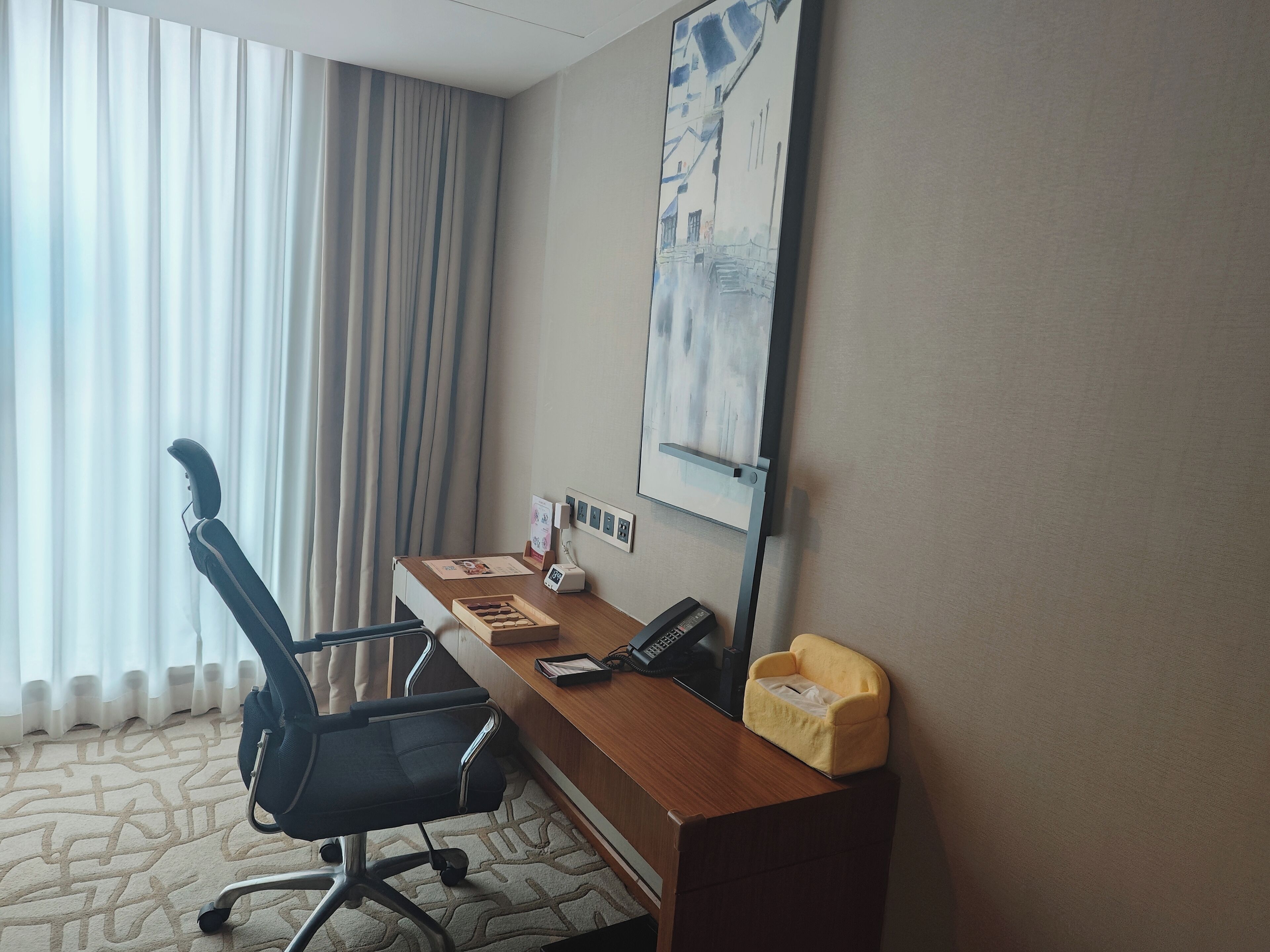 A hotel room with a desk, chair, telephone, and a painting on the wall.