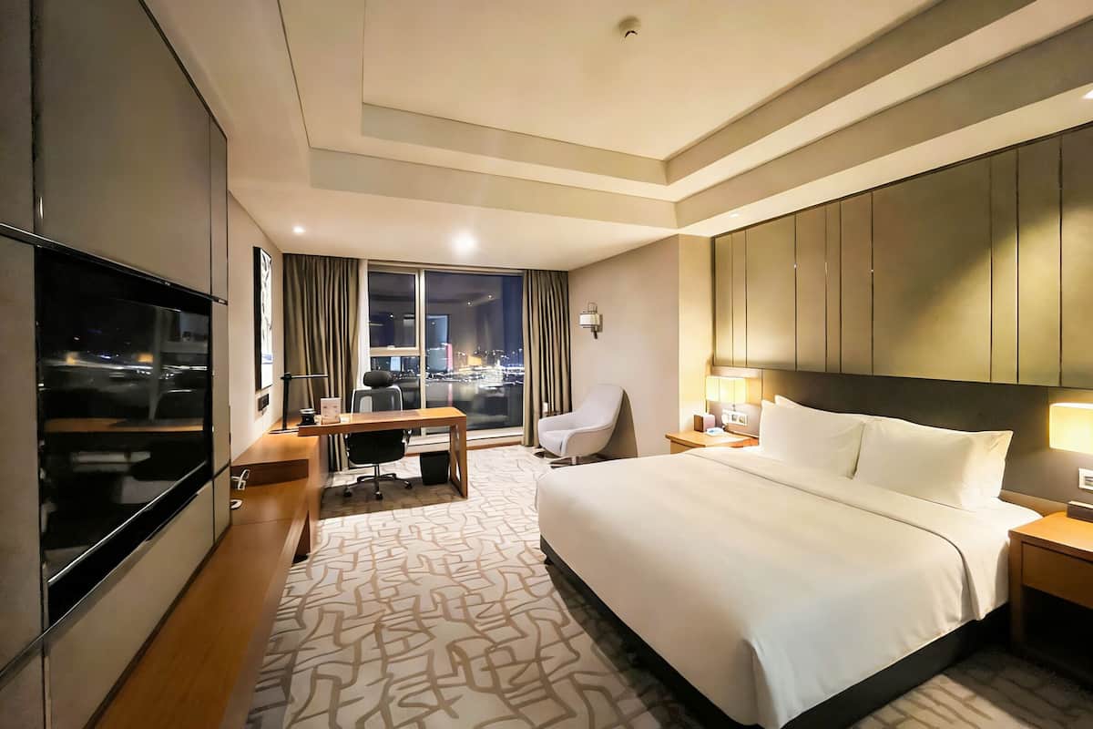 Ein Hotelzimmer mit einem großen Bett, einem Schreibtisch, einem Sessel, einem Fernseher und Blick auf die beleuchtete Stadt.