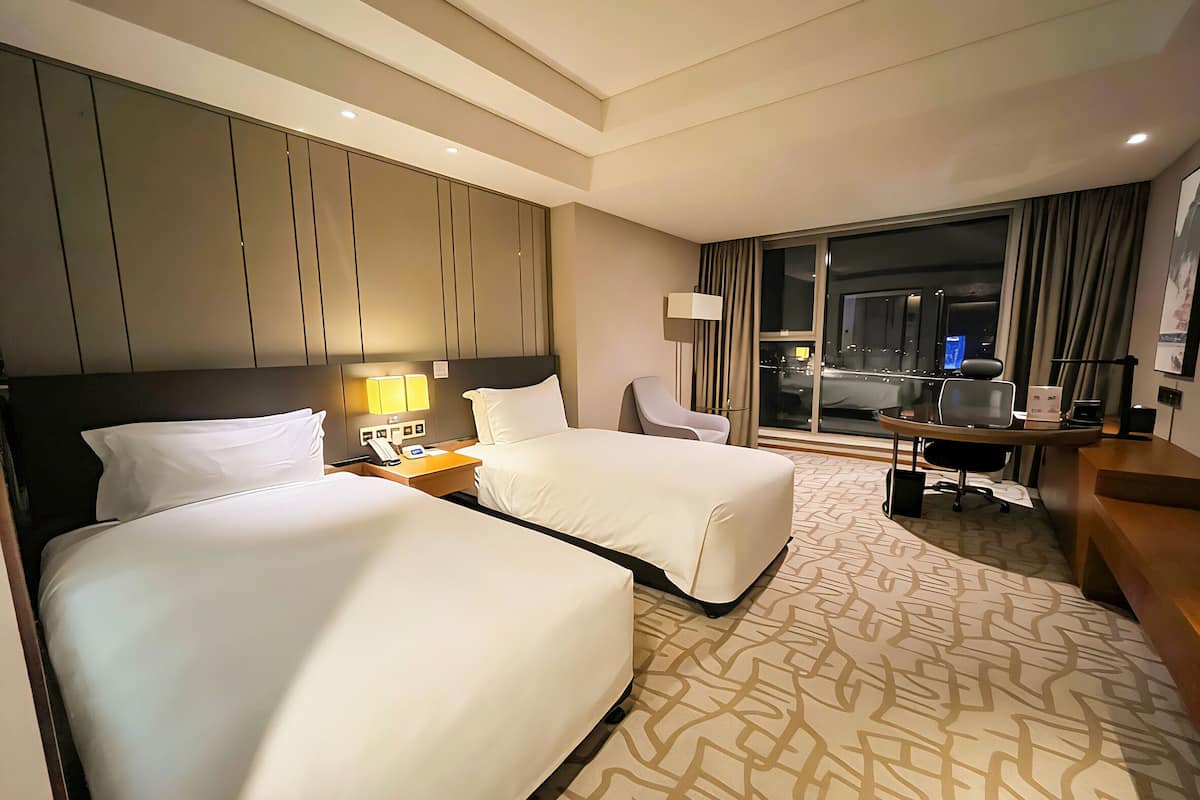 Ein Hotelzimmer mit zwei Betten, einem Schreibtisch, einem Stuhl, einer Lampe und einem großen Fenster mit Blick auf die Stadt.