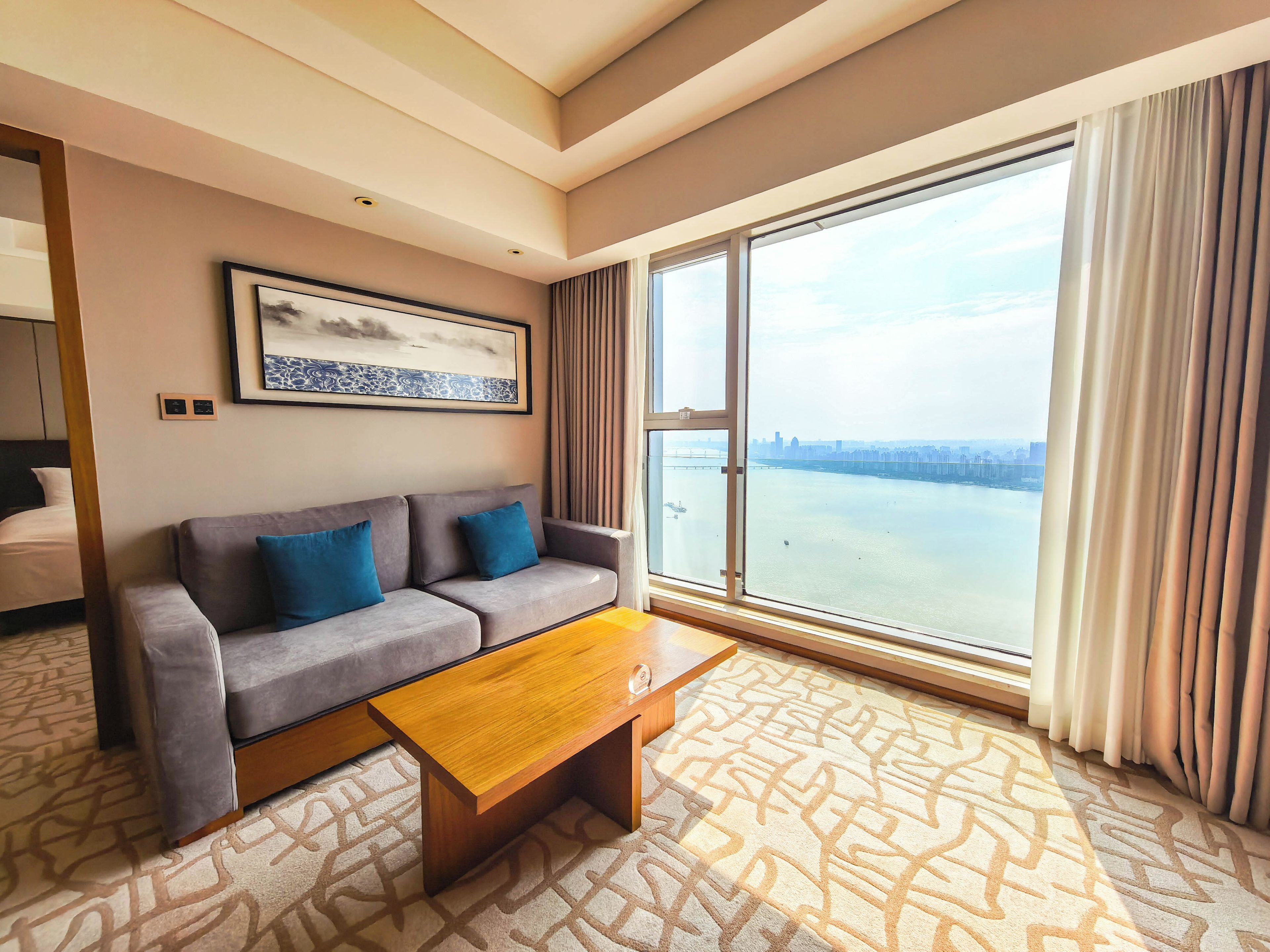 A hotel room with a sofa, a coffee table, a large window with a city view, and a framed picture on the wall.