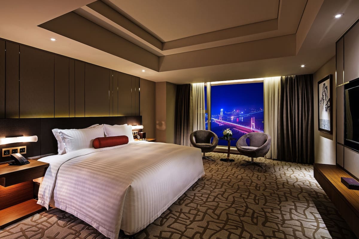 Ein Hotelzimmer mit einem großen Bett, einem Schreibtisch mit Telefon, einem Sessel, einem Fernseher und Blick auf eine nächtliche Skyline.