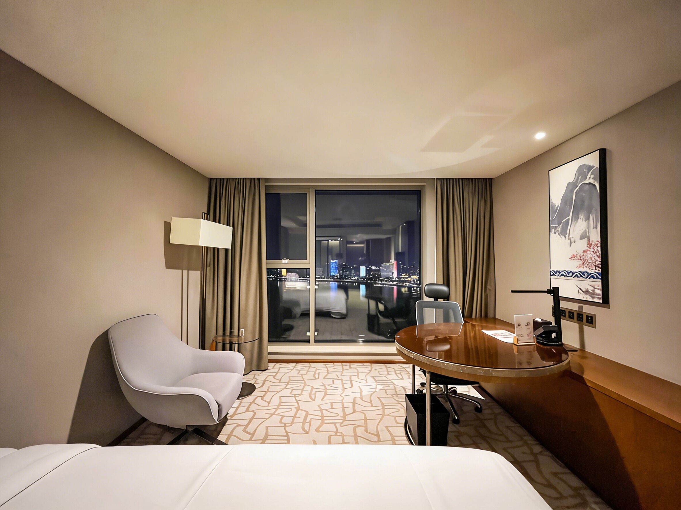 Habitación de hotel con cama, escritorio, silla, lámpara, ventana grande con vistas a la ciudad y un cuadro en la pared.