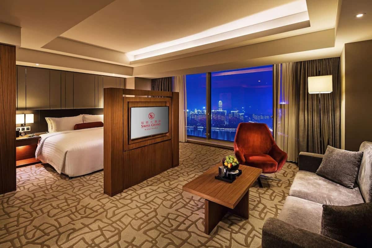 Ein Hotelzimmer mit einem Bett, einem Schreibtisch, einem roten Sessel, einer Couch und einem großen Fenster mit Blick auf die Stadt.