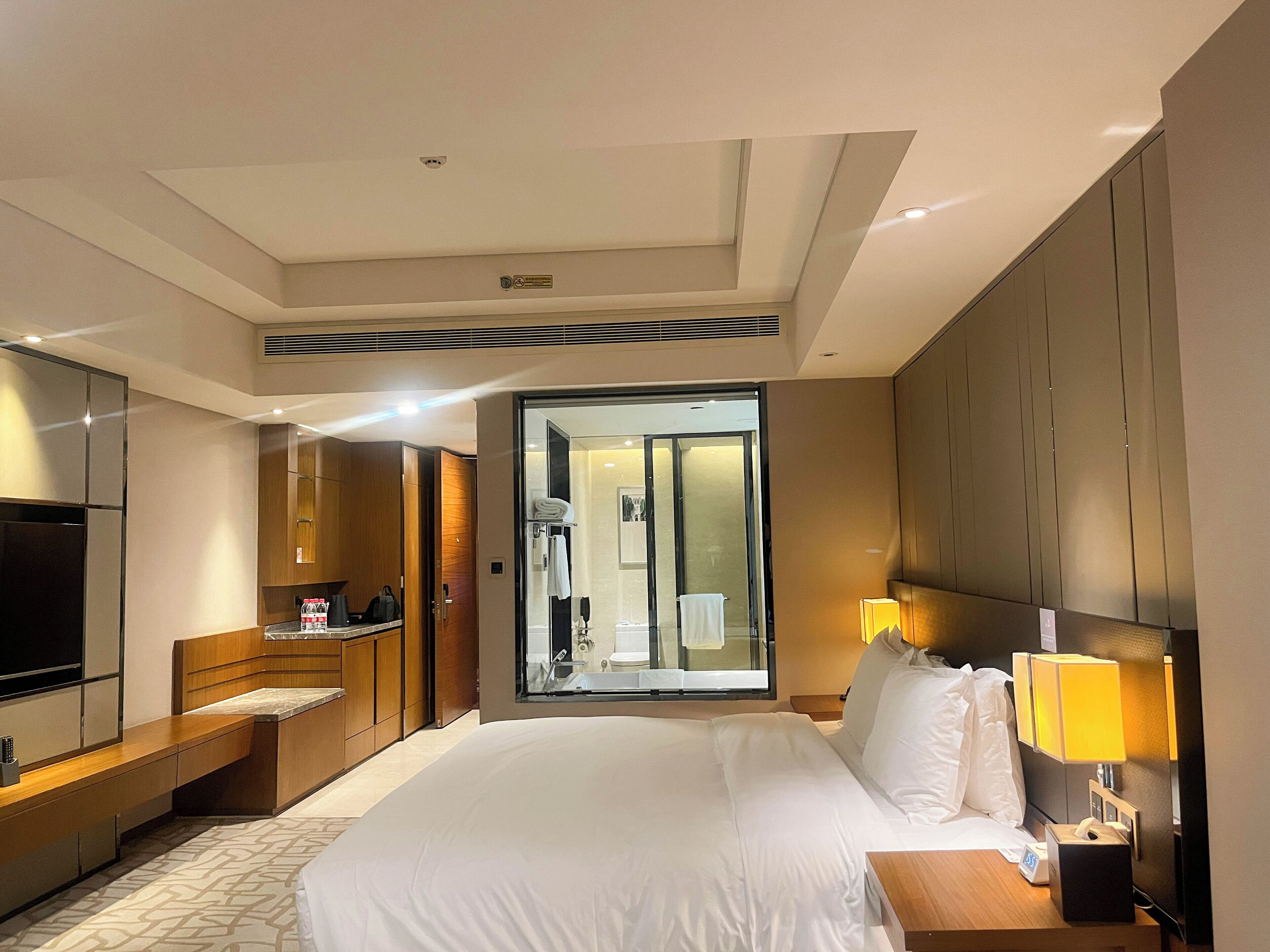 A hotel room with a large bed, a TV, a desk with a lamp, and a bathroom visible through a glass door.