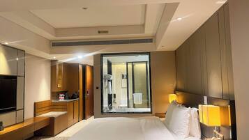 A hotel room with a large bed, a TV, a desk with a lamp, and a bathroom visible through a glass door.