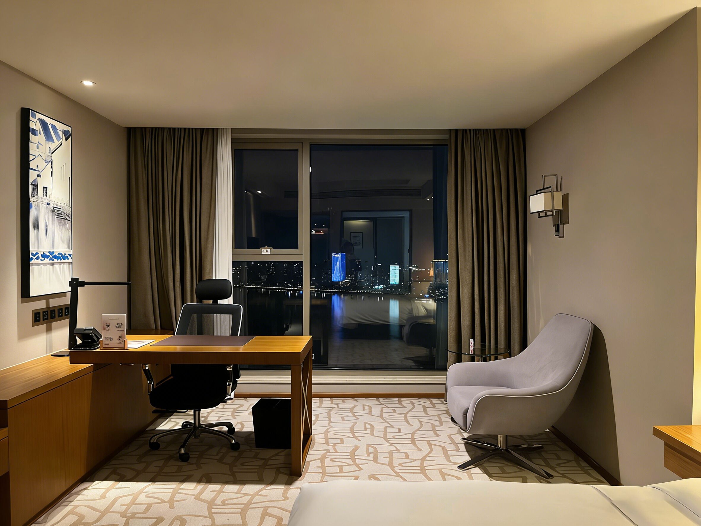 A hotel room with a desk, chair, and bed. It features a city view through large windows.
