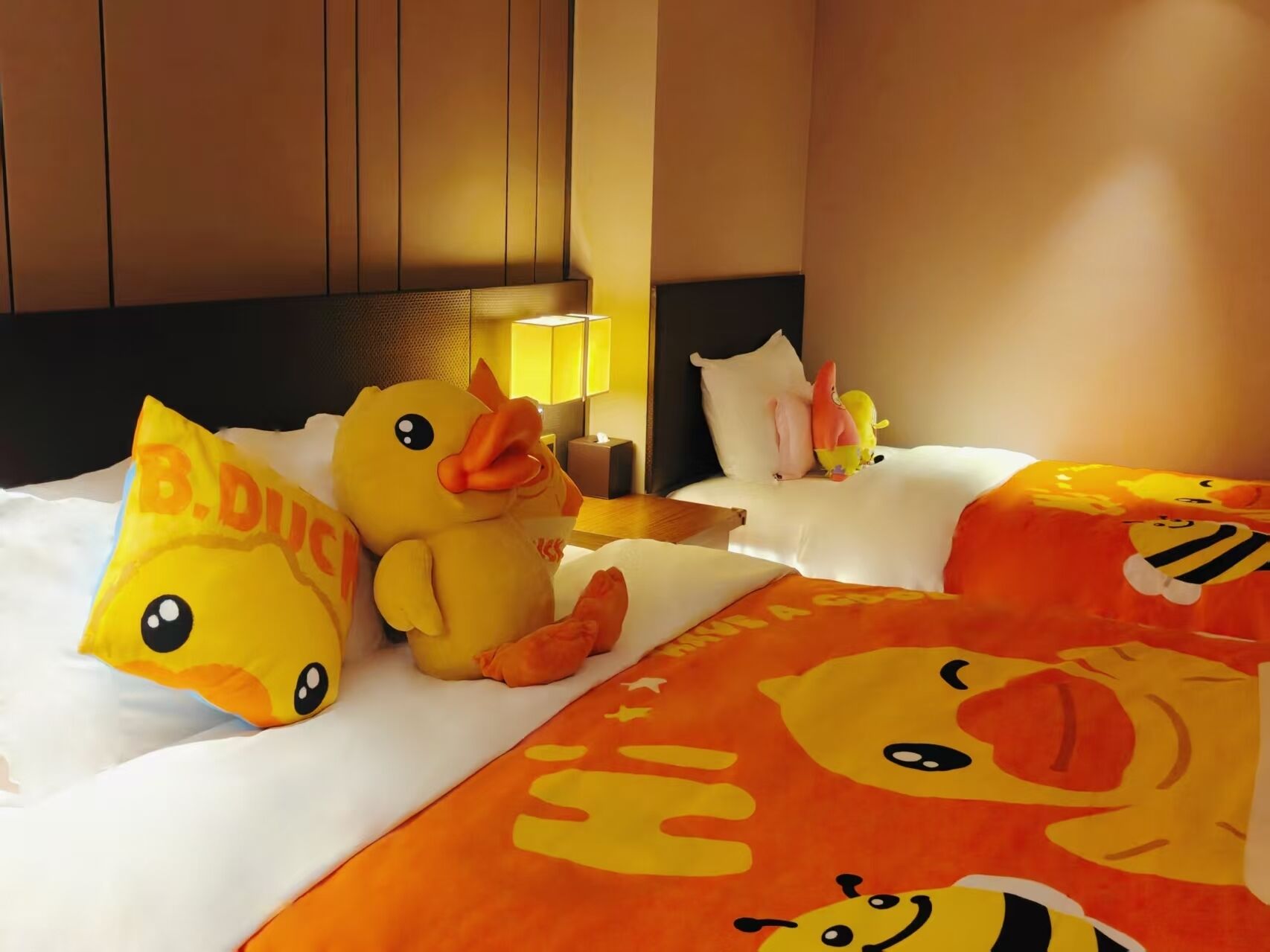 A hotel room with two beds, each adorned with plush duck pillows and a blanket featuring cartoon duck designs.